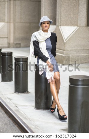 Dressing in a cropped jacket, a fitted dress, open toes shoes and a long scarf, wearing a woolen Fedora hat, a young black woman is sitting on a metal column outside a building, relaxing.