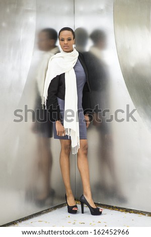 Dressing in a woolen cropped jacket, a fitted dress, a long scarf and high heel open toes shoes, a young black fashion girl is standing by a metal structure, waiting for you.