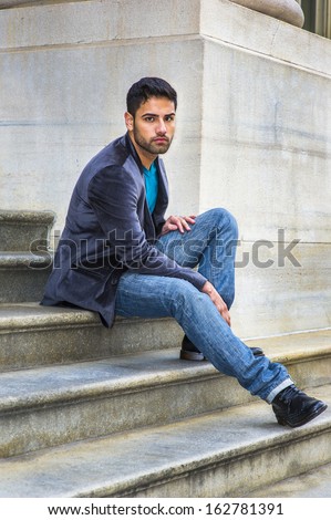 Dressing in a dark purple woolen blazer,  blue jeans and black leather shoes, a young handsome guy with a little bread and mustache is sitting on steps outside an office, thoughtfully looking at you.