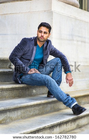 Dressing in a dark purple woolen blazer,  blue jeans and black leather shoes, a young handsome Middle Eastern guy with bread and mustache is sitting on steps outside an office, looking around.