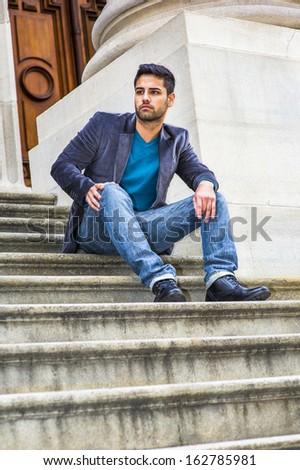 Dressing in a dark purple woolen blazer,  blue jeans and black leather shoes, a young Middle Eastern guy with bread and mustache is sitting on steps in the front doorway, relaxing and thinking.