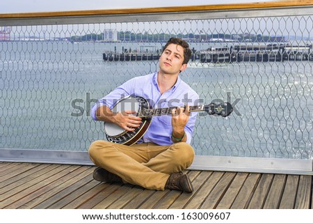 Against a fence, looking up, crossing legs  and thinking,  a young musician is sitting on the deck, playing a banjo / Thinking about You