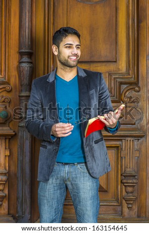 Dressing in a blue woolen blazer, knit sweater and jeans, holding glasses and a red book,  a young handsome professor with bread and mustache is standing in the doorway, smilingly looking forward.