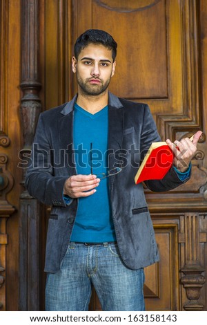 Dressing in a woolen blazer, knit sweater and jeans, holding glasses and a red book,  a young handsome professor with bread and mustache is standing in the office doorway, seriously looking at you.
