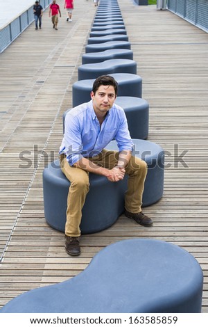 Dressing in a light blue shirt, dark yellow jeans and brown boot shoes, a young handsome guy is sitting on a modern style bench, charmingly looking at you. / Relaxing and Thinking Outside