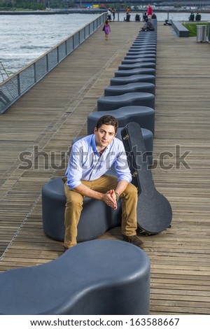 Dressing in a light blue shirt, dark yellow jeans and brown boot shoes, a young handsome musician with a music instrument box is sitting on a modern style bench in the evening,  looking at you.