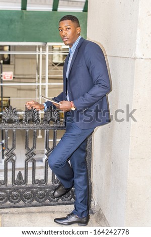Dressing in a blue suit and leather shoes, holding a tablet computer, a young black businessman is standing on the corner outside, looking at you. / Waiting for You.
