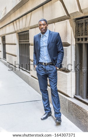 A young black businessman is standing outside by a window on a street and taking a break / Portrait of Young Black Businessman