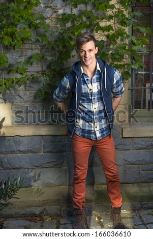 Dressing in a blue and white pattern shirt,  a blue hoody vest,  red jeans, a young guy is standing by a window with green ivy leaves. / Portrait of Young Guy
