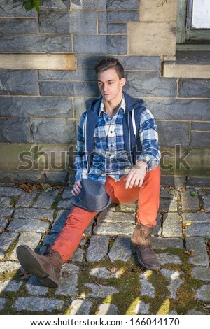 Dressing in blue, white pattern shirt, a hoody vest,  red jeans and brown leather boot shoes, holding a woolen Fedora hat on his knee, a young guy is sitting on the ground into deeply thinking.