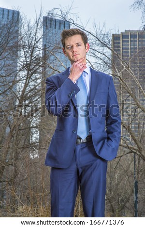 Dressing a blue suit and tie, one hand touching his chin , a young businessman is standing outside and thinking. The background is high business buildings. / Thinking Outside