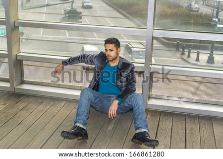 Against a glass wall, a young guy with beard and mustache is sitting on the floor into deeply thinking. Background is over a street. / Cross Road