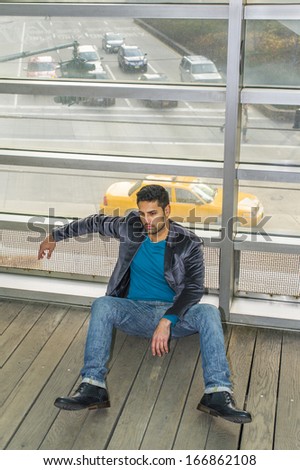 Against a glass wall, a young guy with beard and mustache is sitting on the floor into deeply thinking. Background is over a street with many cars / City Life