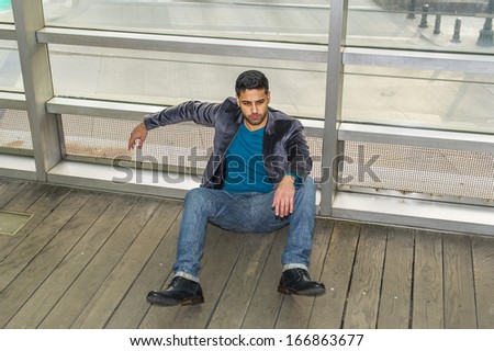 Against a glass wall, a young guy with beard and mustache is sitting on the floor into deeply thinking. Background is over a street. / Frustrated Man