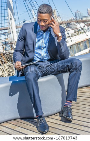 Looking at a tablet computer on his lap, a young black businessman is sitting on a deck and smilingly talking on the phone. The background is a harbor. / Working Outside