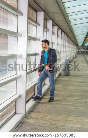 Wearing glasses, holding a red book and a white flower,  a young guy with beard and mustache is standing on the walk way against the glass wall, seriously looking outside. / Waiting for You
