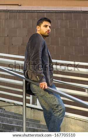 Dressing in dark purple woolen blazer and jeans, a young guy with beard and mustache is standing against a metal railing on stairs, under sunshine of sunset, thinking. / Thinking Outside