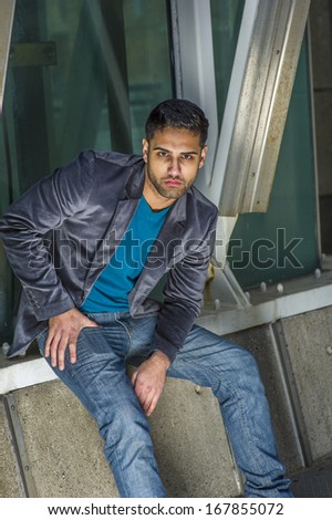 Dressing in a dark purple woolen blazer, a blue sweater and jeans , a young handsome guy with beard and mustache is sitting on the corner by a structure, bending over, thoughtfully looking at you.