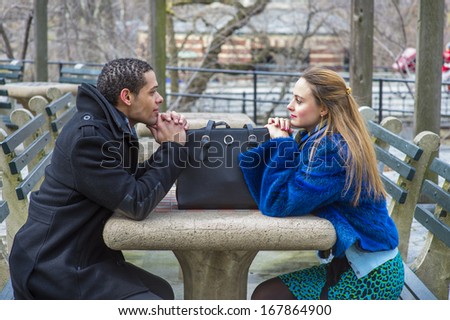 A girl dressing in a blue faux fur plus size jacket and  a guy dressing in a black pea coat are sitting crossing a table, looking at each other, with a leather bag. / Two Friends and One Bag
