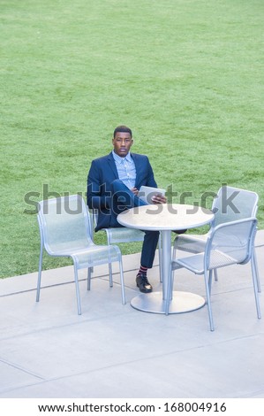 A young handsome black man is sitting on a chair by a green lawn and reading at a small computer. / Reading Outside