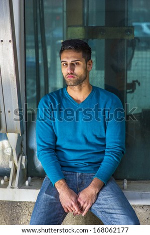Dressing in a blue sweater and jeans , a young handsome guy with beard and mustache is sitting on the corner by a structure, thoughtfully looking at you. / Tough Time