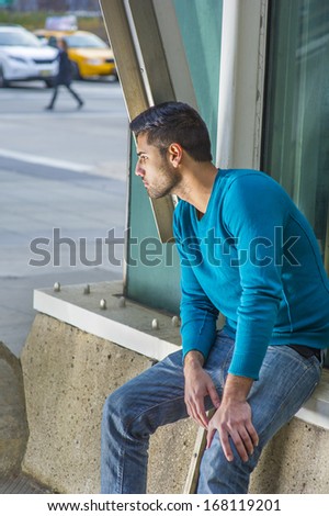 Dressing in a blue sweater and jeans , a young handsome guy with beard and mustache is sitting on the corner of the street, bending over, looking down, lost in thought. / City Life