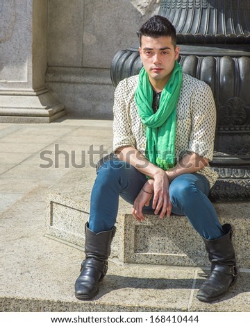 Dressing in a patterned shirt,  long green scarf, blue pants, long leather boots,  a young handsome guy is sitting there and relaxing. / Traveler