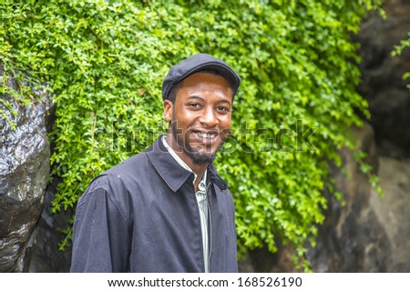 Dressing in a waterproof jacket, a ivy cap,  a young handsome black guy is standing by rocks with green plants in a raining day, smilingly looking at you. / Raining Day