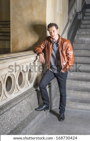 Dressing in a brown leather jacket, black pants, one hand putting in a pocket,  a young handsome guy is standing by railing under stairs, looking at you. / Portrait of Young Guy
