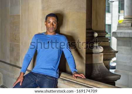 Dressing in a light blue, long sleeve T shirt, a young handsome black student is standing at a balcony against a wall, sincerely looking at you. / Portrait of Young Black Guy