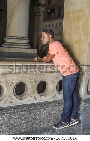 Dressing in a light orange short sleeve shirt, jeans and leather sneakers,  a young handsome black guy is tiptoeing and bending over a railing at a balcony, looking down and watching. / Observation
