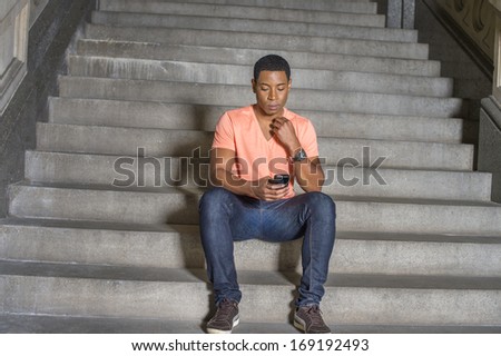 Dressing in a light orange short sleeve V neck shirt, jeans,  a young handsome black guy is sitting on stairs, checking messages on his mobile phone. / Text