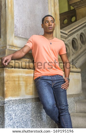 Dressing in a light orange short sleeve V neck shirt, jeans,  a young handsome black student is standing downstairs against a column, looking forward. / Waiting for You