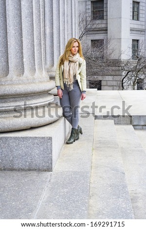 Dressing in a light green jacket, long beige scarf, tight gray fashion pants and green boots, a young pretty woman is standing by a column outside an office building / Portrait of Businesswoman