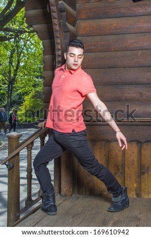 Dressing in a red Polo shirt, black pants and leather boots, stretching arms and turning over legs,  a young guy is standing by an old fashion style, wooden house doorway in morning, exercising.