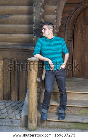 Dressing in a green striped long sleeves Henley shirt, black pants and leather boots, a young handsome guy is standing outside a wooden house and taking a break. / Waiting for You