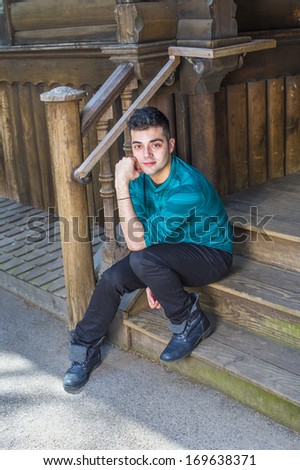 Dressing in a green striped long sleeves Henley shirt, black pants and leather boots, a young handsome guy is sitting on stairs outside a wooden house and taking a break. / Relaxing Outside