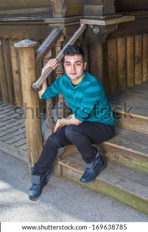 Dressing in a green striped long sleeves Henley shirt, black pants and leather boots, a young handsome guy is sitting on stairs outside a wooden house and taking a break. / Relaxing Outside