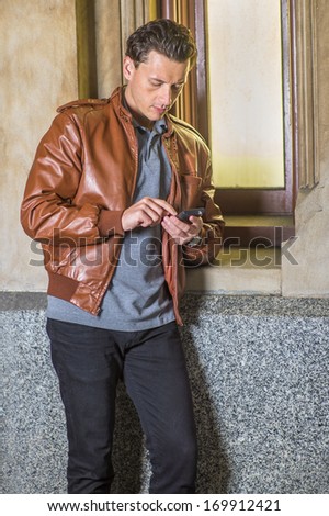 Dressing in a brown leather jacket, black pants,  a young handsome guy is standing by a small window, checking messages on his cell phone. / Text