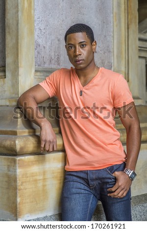 Dressing in a light orange short sleeve V neck shirt, jeans,  a young handsome black student is standing downstairs by a column, confidently looking at you. / Portrait of Young Black Student