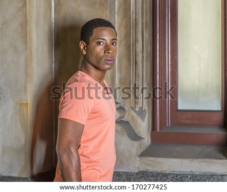 Dressing in a light orange short sleeve V neck shirt, jeans,  a young handsome black student is standing by a window, looking around and staring you. / Portrait of Young Black Student