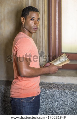 A young handsome black student is standing by a window, reading a book. / Reading Outside