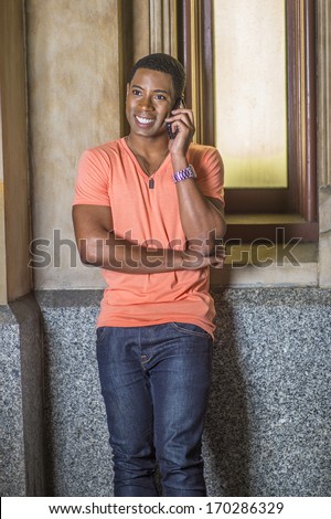 Dressing in a light orange short sleeve V neck shirt, jeans,  a young handsome black student is standing by a small window, smiling and talking on the phone. / Making a Phone Call