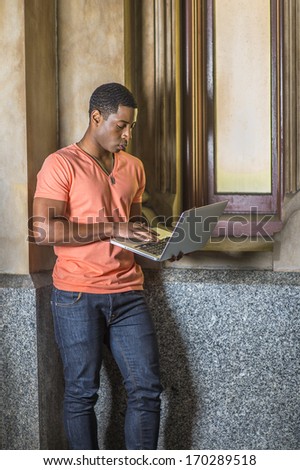 Dressing in a light orange short sleeve V neck shirt, jeans, a young handsome black student is standing by a small window, working on a laptop computer. / Study Outside
