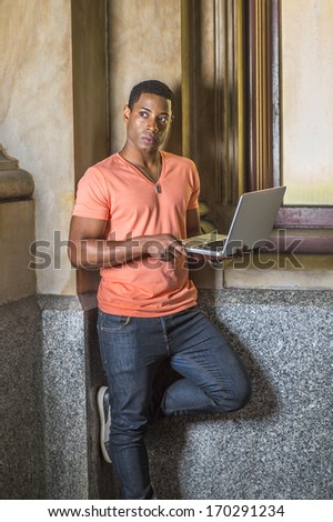 Dressing in a light orange short sleeve V neck shirt, jeans, a young handsome black student is standing by a window, working on a laptop computer. / Study Outside