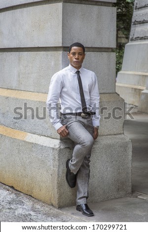 Dressing in white shirt, a black tie, gray pants and leather shoes, a young handsome black college student is standing by a stone wall, confidently looking at you. / Portrait of a College Student