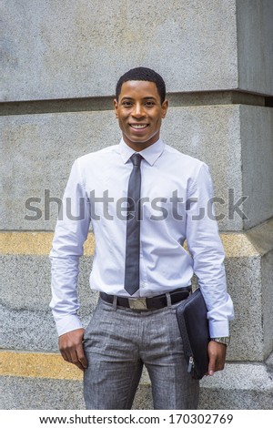 Dressing in white shirt, a black tie, gray pants, carrying a briefcase, a young handsome black college student is standing by a stone wall, smilingly looking at you. / Portrait of a College Student