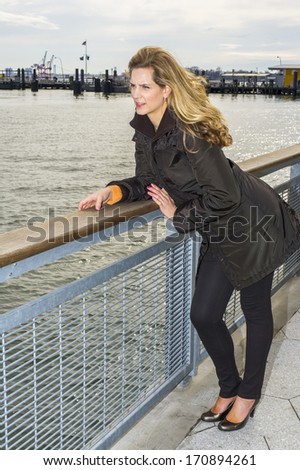 A pretty woman is standing and bending over a fence by a river in a harbor, looking forward / Waiting for You
