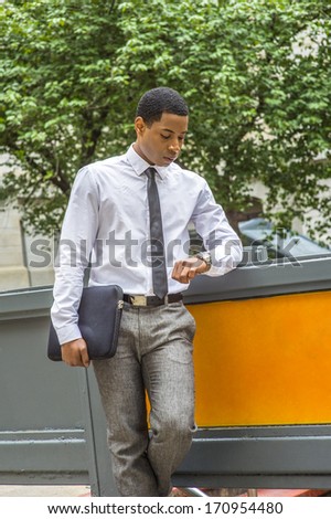 Dressing in white shirt, a black tie, gray pants, and carrying a black briefcase, a young handsome black businessman is looking at his watch and waiting for you. / Time