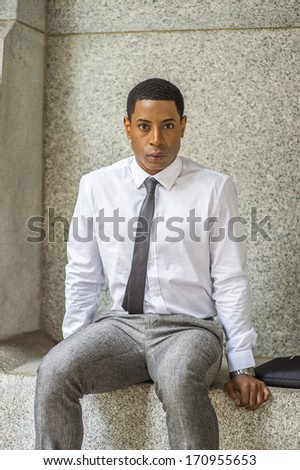 Dressing in white shirt, a black tie, gray pants, and carrying a black briefcase, a young handsome black businessman is sitting on a stone bench, waiting outside / Waiting